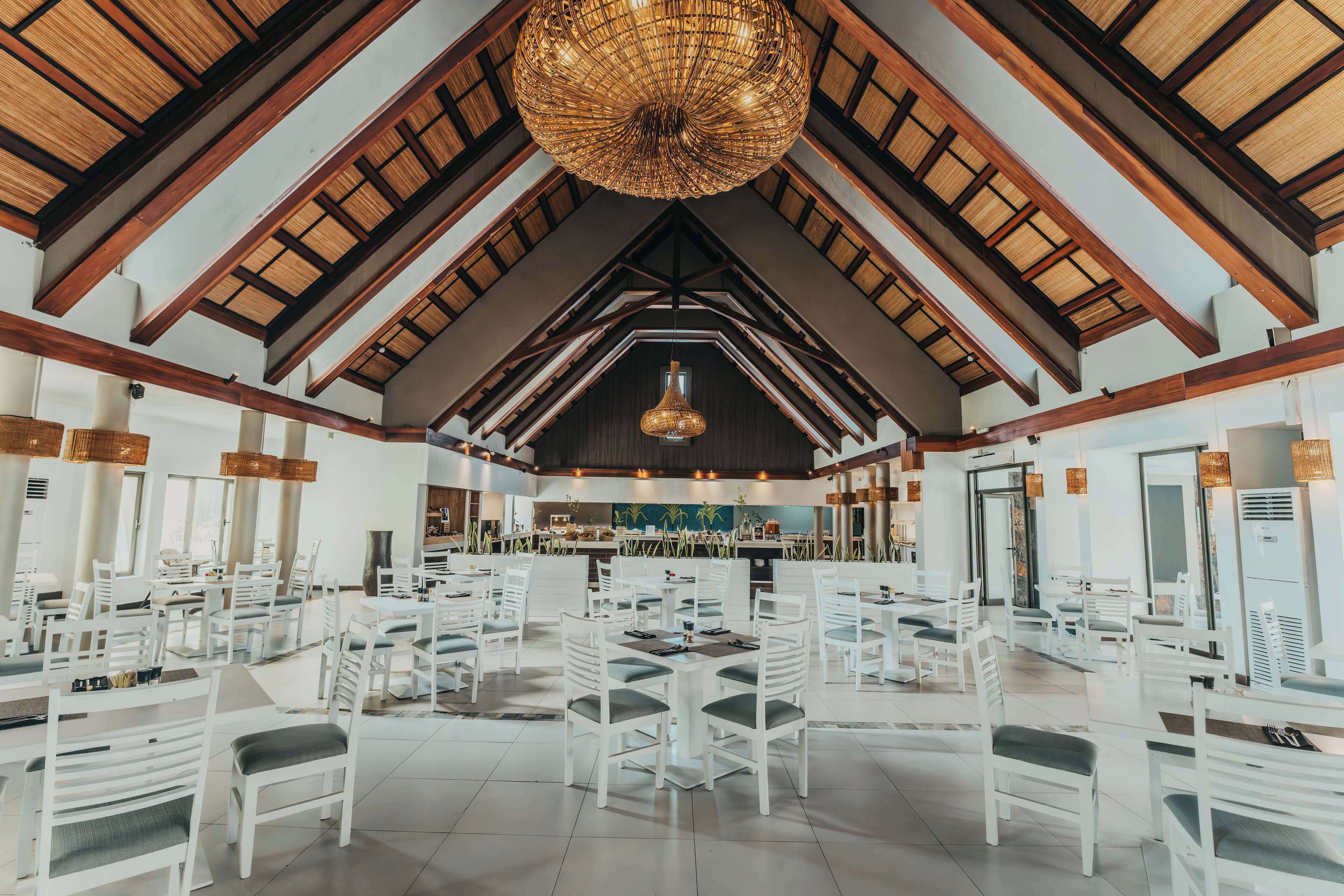 Une salle à manger spacieuse avec des tables et des chaises blanches, un grand lustre et une cuisine en arrière-plan.