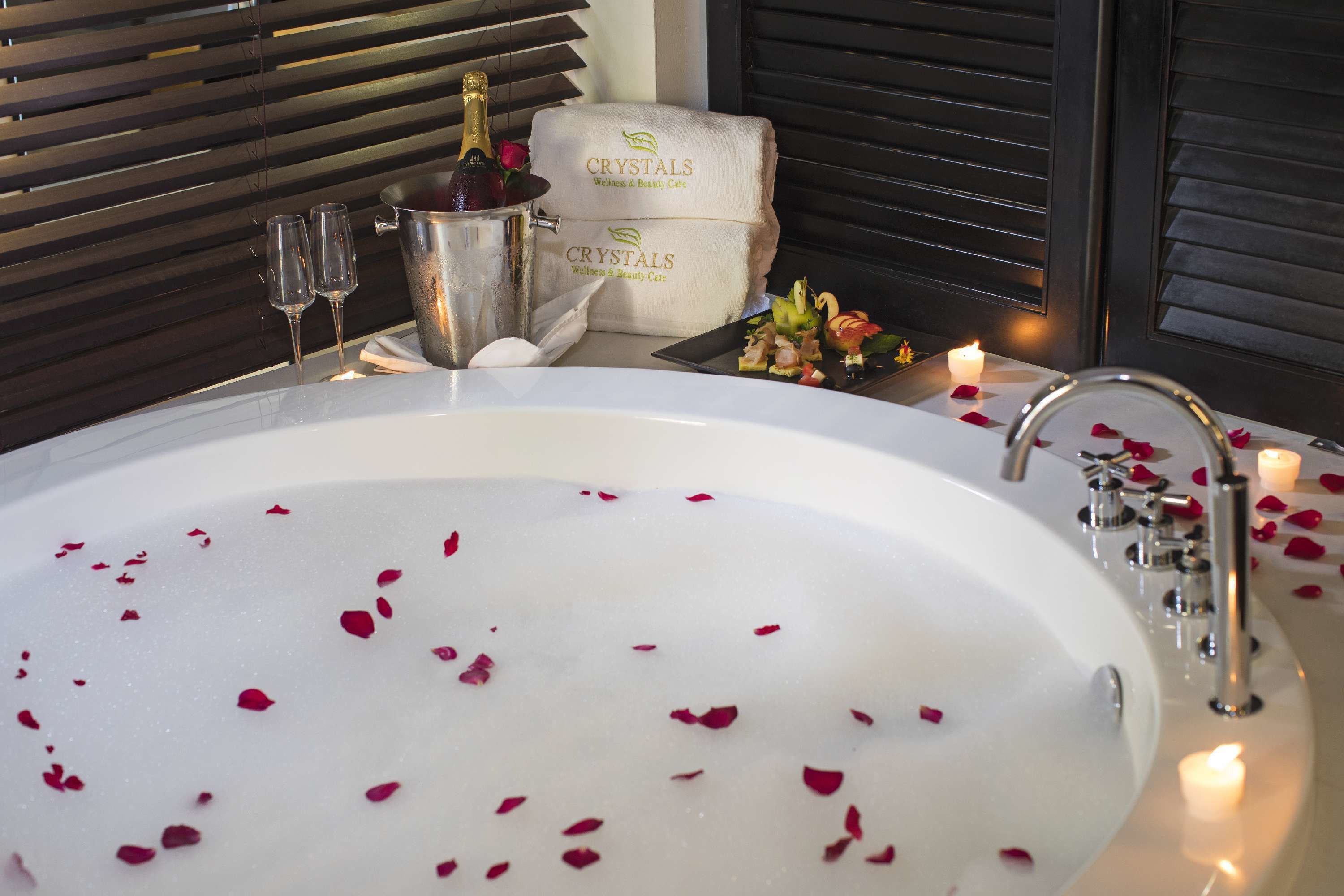 A bathtub filled with water and rose petals, a champagne bottle, two glasses, and a tray with food and flowers.