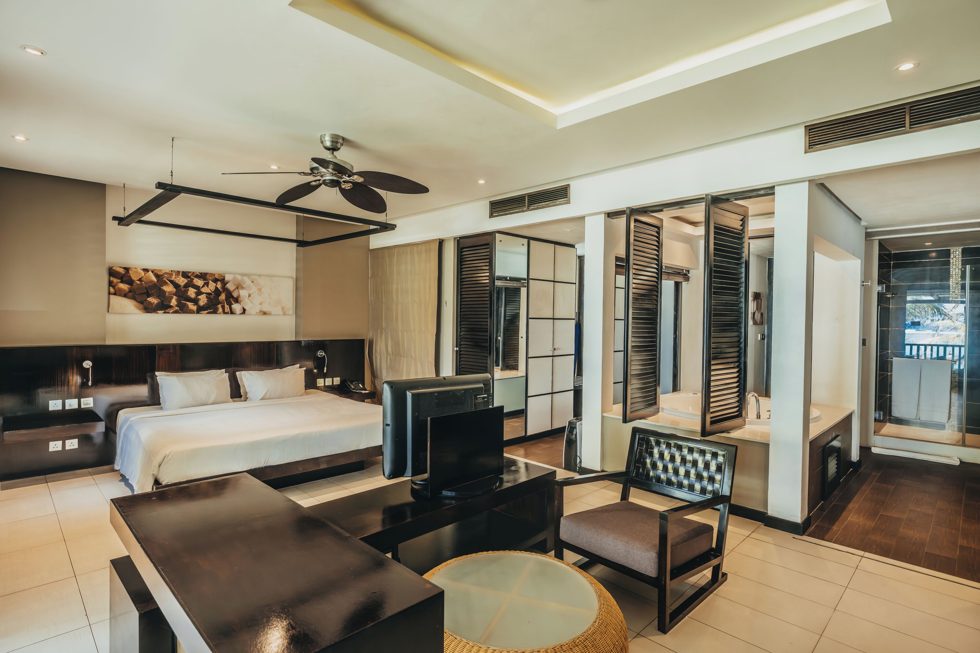 A modern hotel room with a bed, a TV, a desk, and a chair. There is a ceiling fan and a large window.