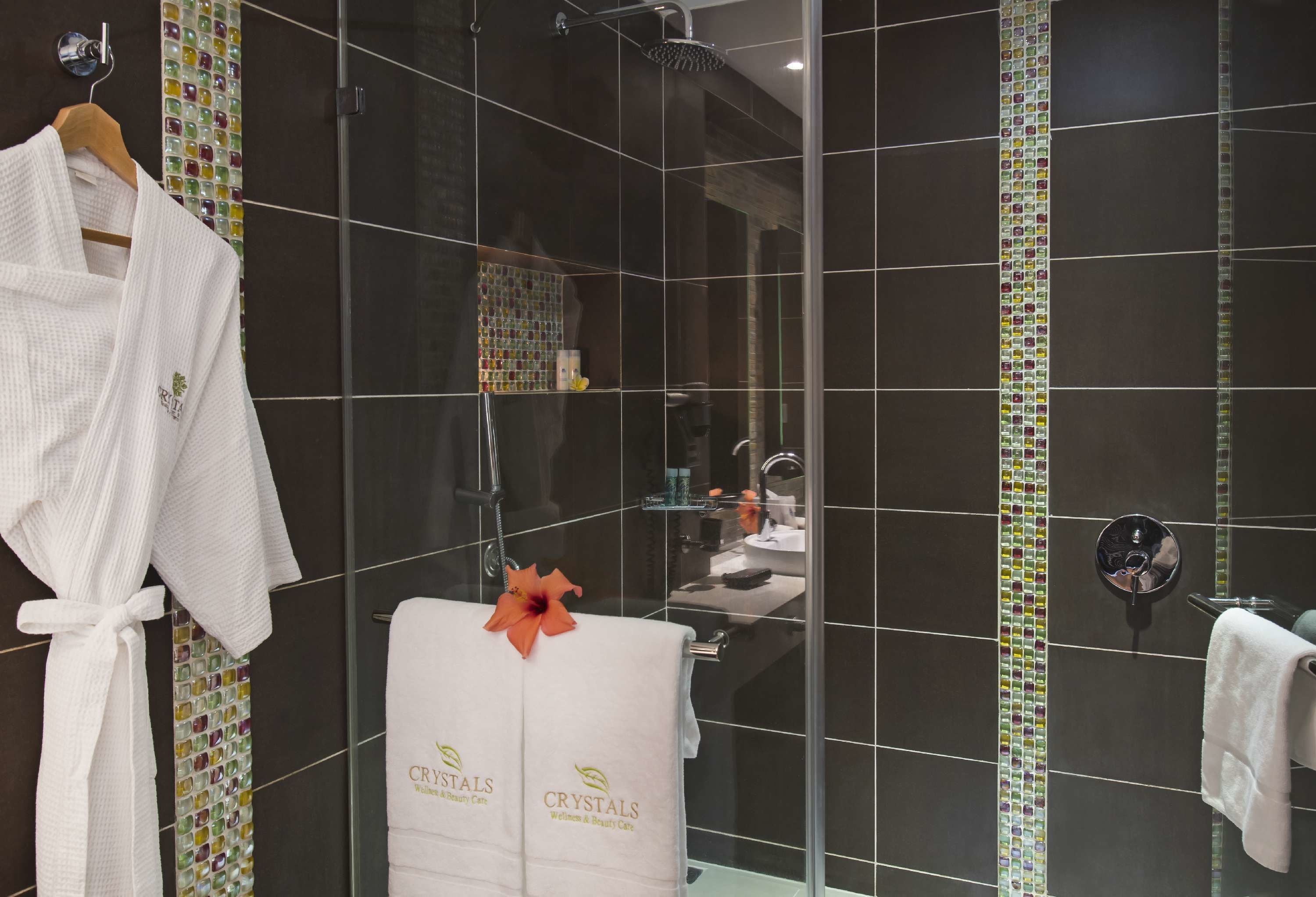 Un baño con baldosas oscuras, mampara de ducha de vidrio, bata blanca y toalla con la marca 'CRYSTALS Wellness & Beauty Care'.