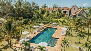 A resort with a rectangular pool surrounded by lounge chairs and umbrellas, set in a tropical garden with palm trees and a clear sky.