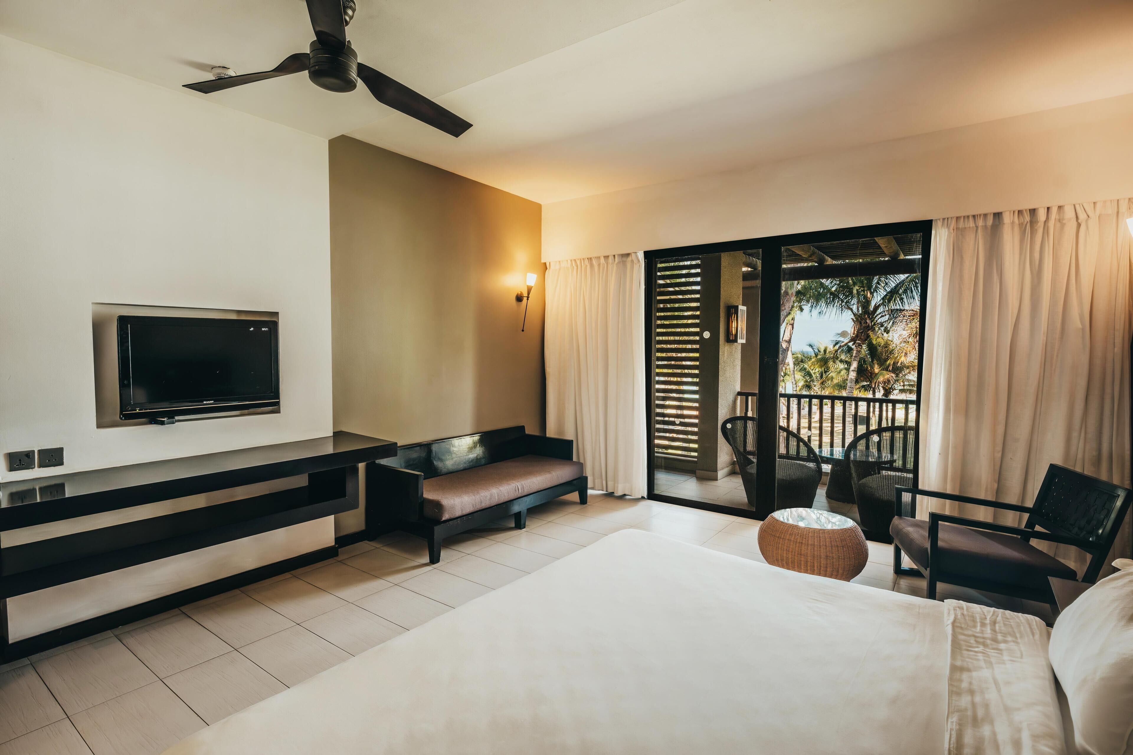 A modern hotel room with a flat-screen TV, a sofa, a bed, a ceiling fan, and a balcony with outdoor seating.