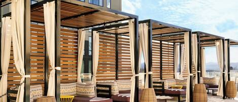 A rooftop lounge area with multiple seating options, including cushioned benches and chairs, under a wooden pergola structure.