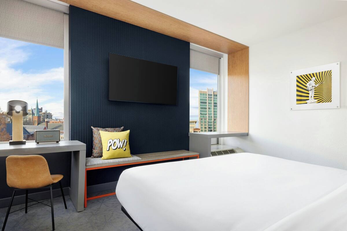 A modern hotel room with a bed, a desk with a chair, a large window with a city view, and a flat-screen TV mounted on the wall.