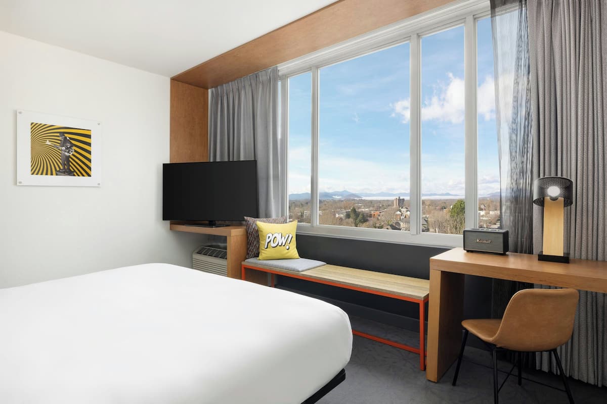 A hotel room with a bed, a desk with a chair, a television, a window with a view of the city, and a pillow with "POW!" on it.