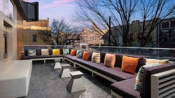 A modern outdoor seating area with geometric patterns on the floor, a long bench with colorful cushions, and a view of buildings and trees in the background.