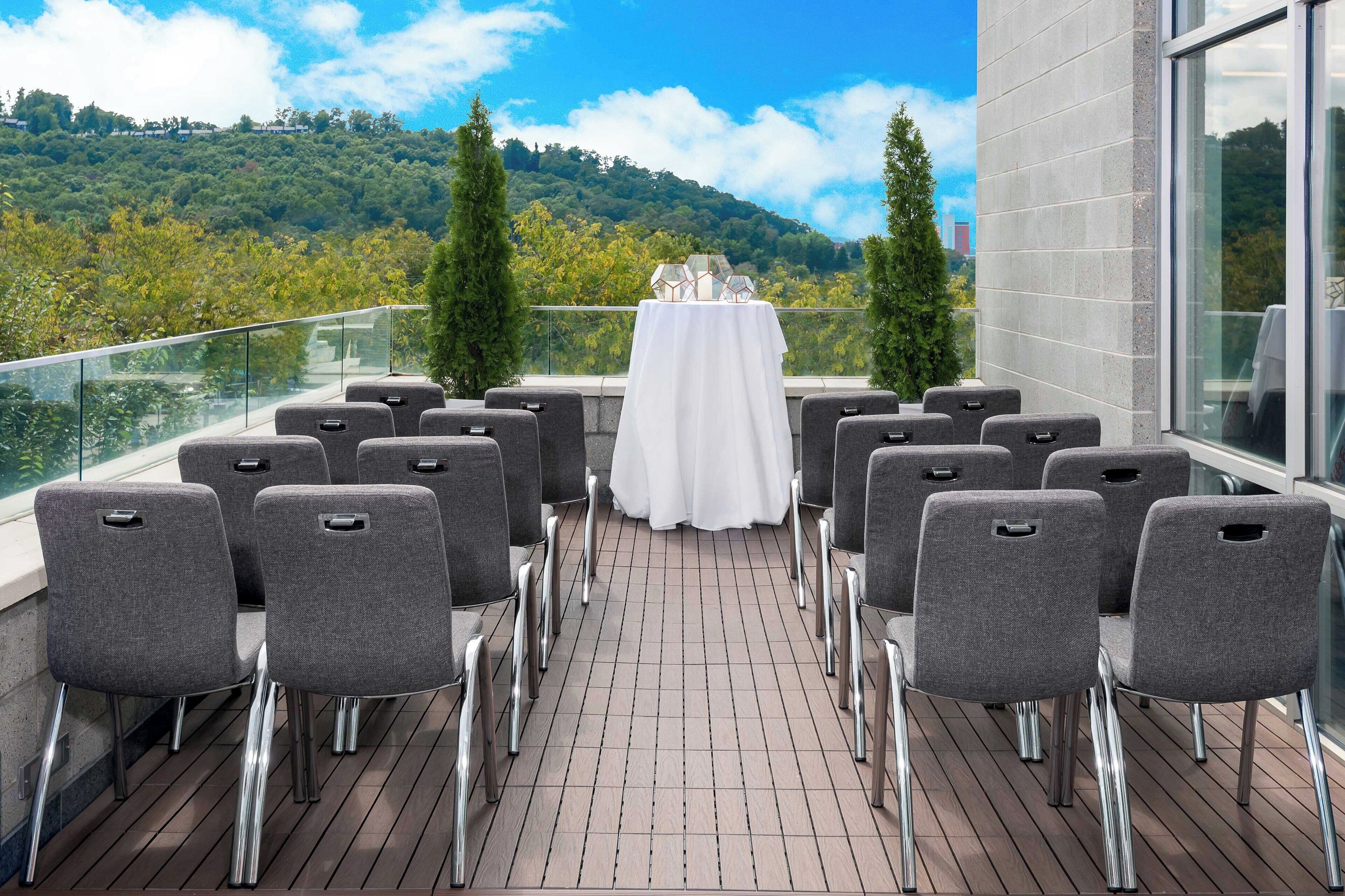 A terrace with grey chairs arranged in rows, a table with a white tablecloth, and a view of a forested hill.