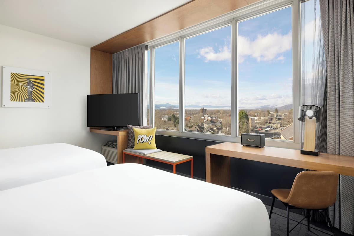 A hotel room with two beds, a desk with a chair, a television, and a view of a cityscape through large windows.