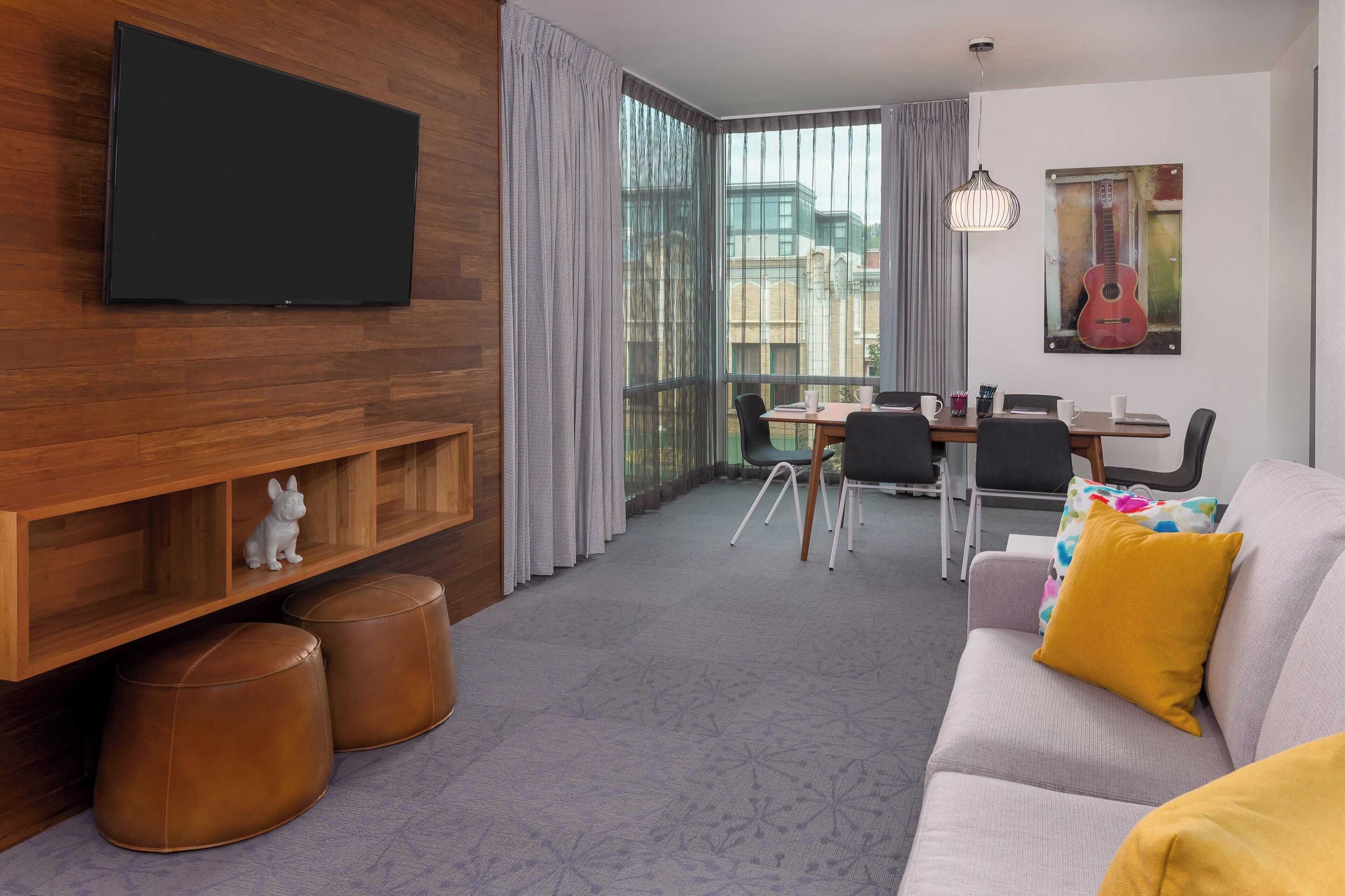 Un salon moderne avec un téléviseur à écran plat, des étagères en bois, des poufs en cuir, un canapé gris avec un coussin jaune, une table à manger avec des chaises et une grande fenêtre avec des rideaux.