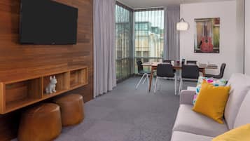 Een moderne woonkamer met een flatscreen televisie, houten schapenkasten, leren poefs, een grijze bank met een geel kussen, een eettafel met stoelen en een groot raam met gordijnen.