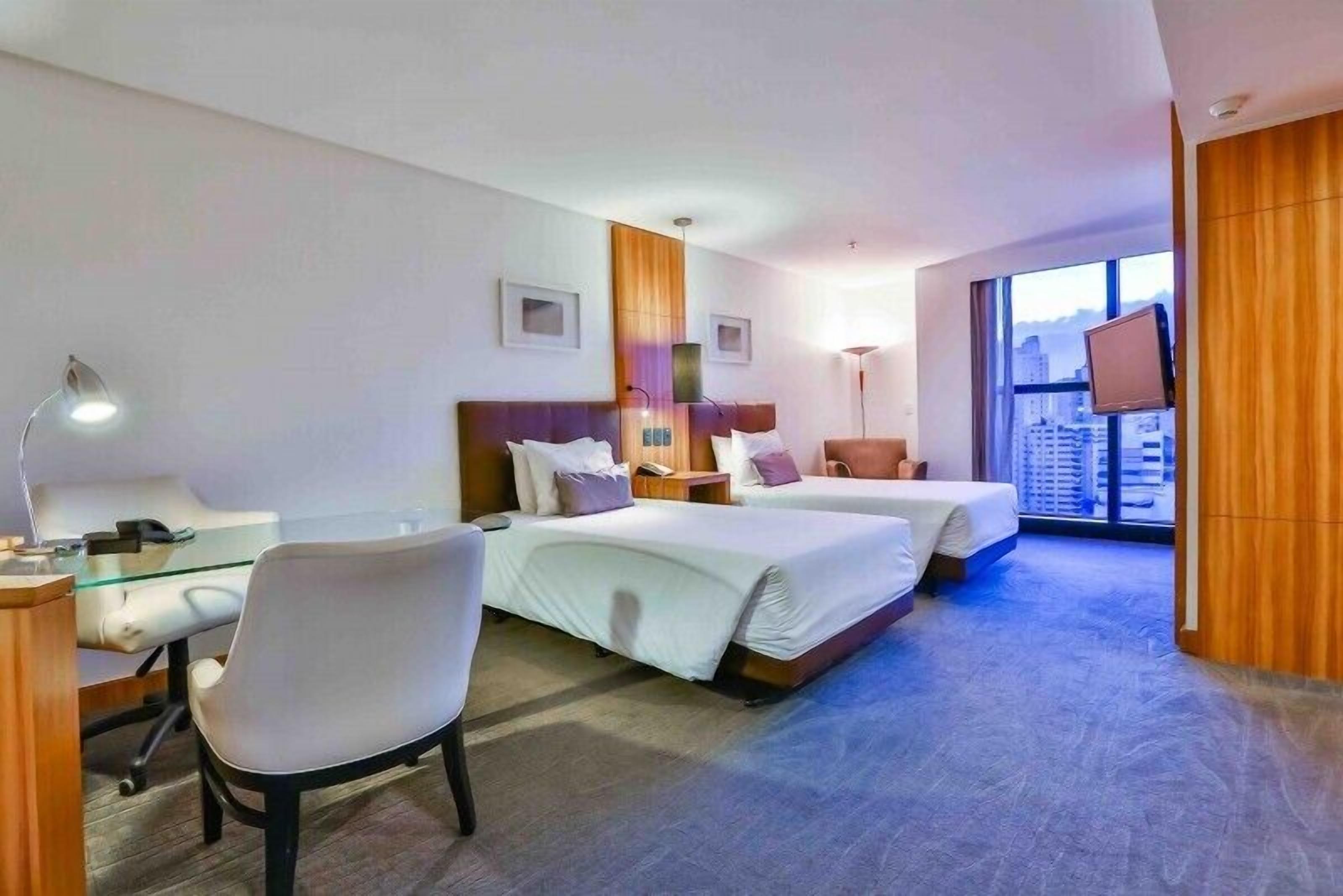 Habitación de hotel con dos camas, un escritorio con silla, televisión y un ventanal con vistas a la ciudad.