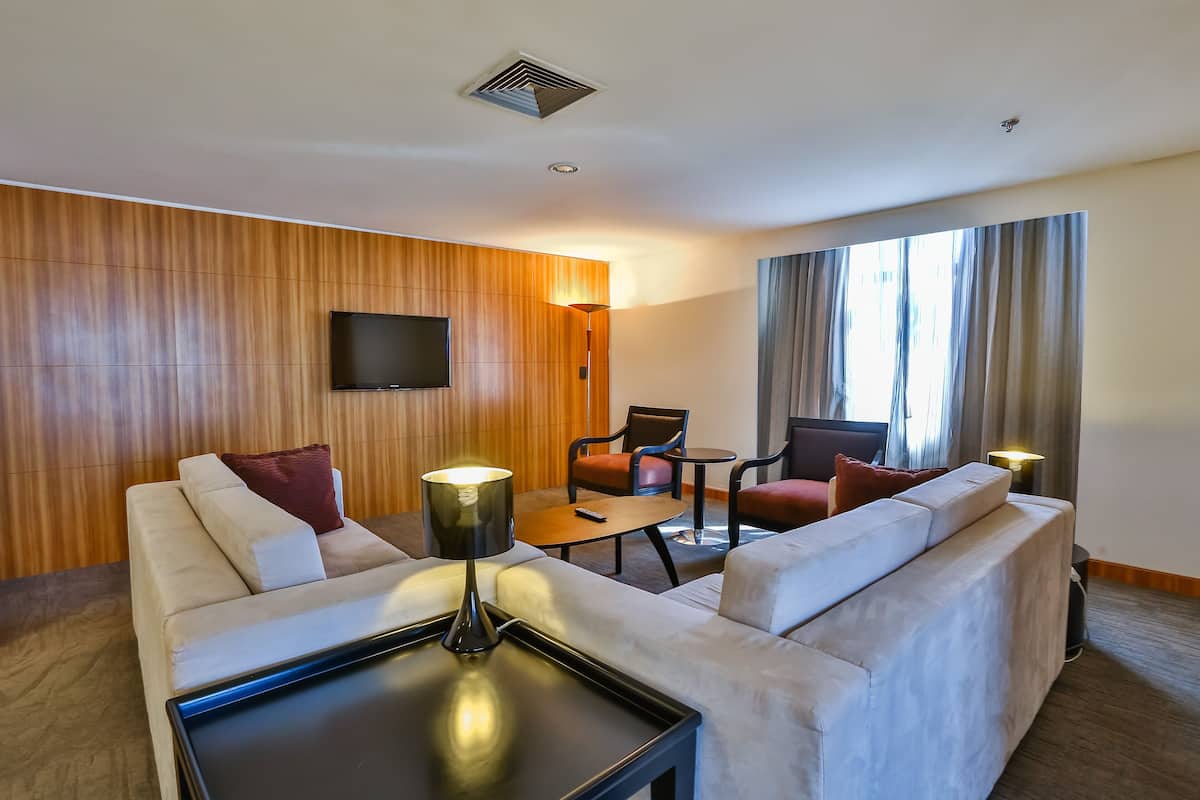 Una habitación de hotel moderna con un conjunto de sofá, una mesita, un televisor montado en la pared y un ventanal con cortinas.