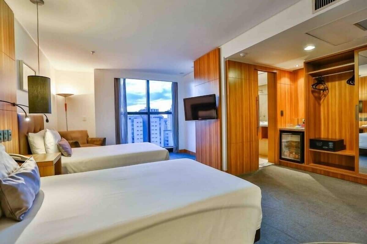 Habitación de hotel con dos camas, televisión, armario y ventana grande con vistas a la ciudad.