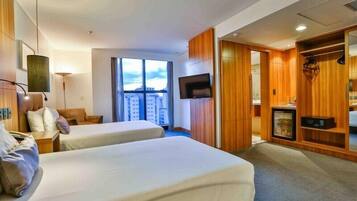 Quarto de hotel com duas camas, televisão, guarda-roupa e uma janela grande com vista para a cidade.