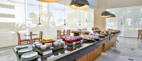 A buffet setup with various food items and plates, overhead lighting, and a view of the outside through large windows.