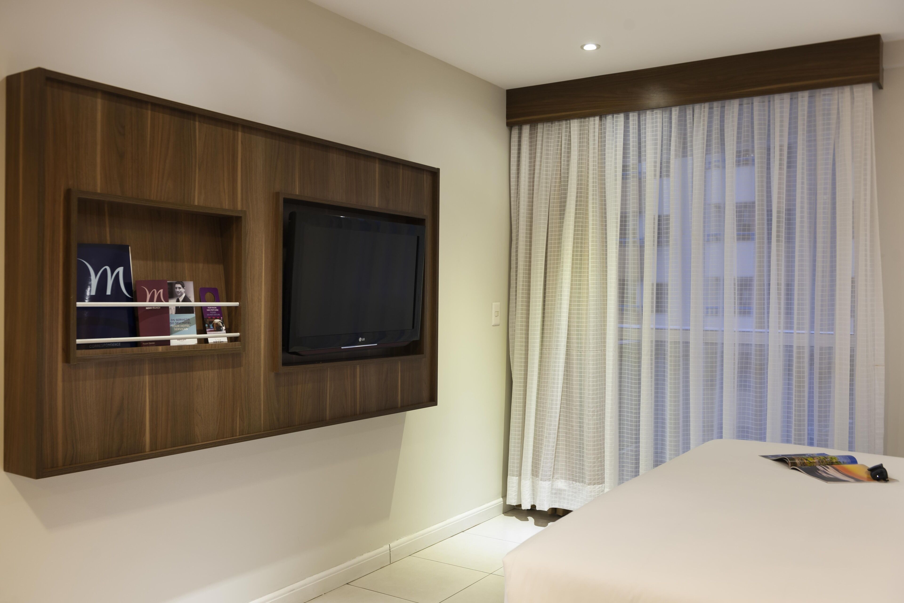 A hotel room with a mounted TV, a bed, and a window with sheer curtains.