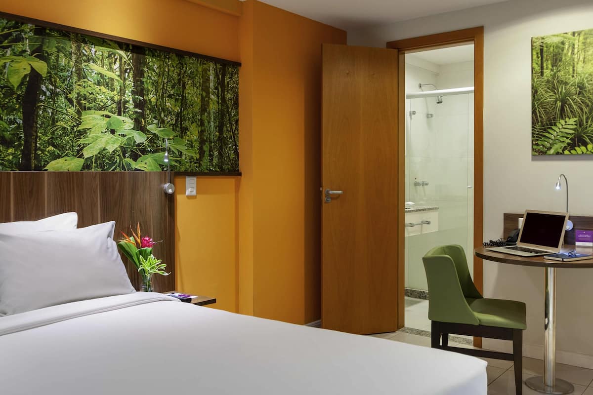 A hotel room with a bed, a desk with a laptop, a chair, a shower, and a large wall mural.