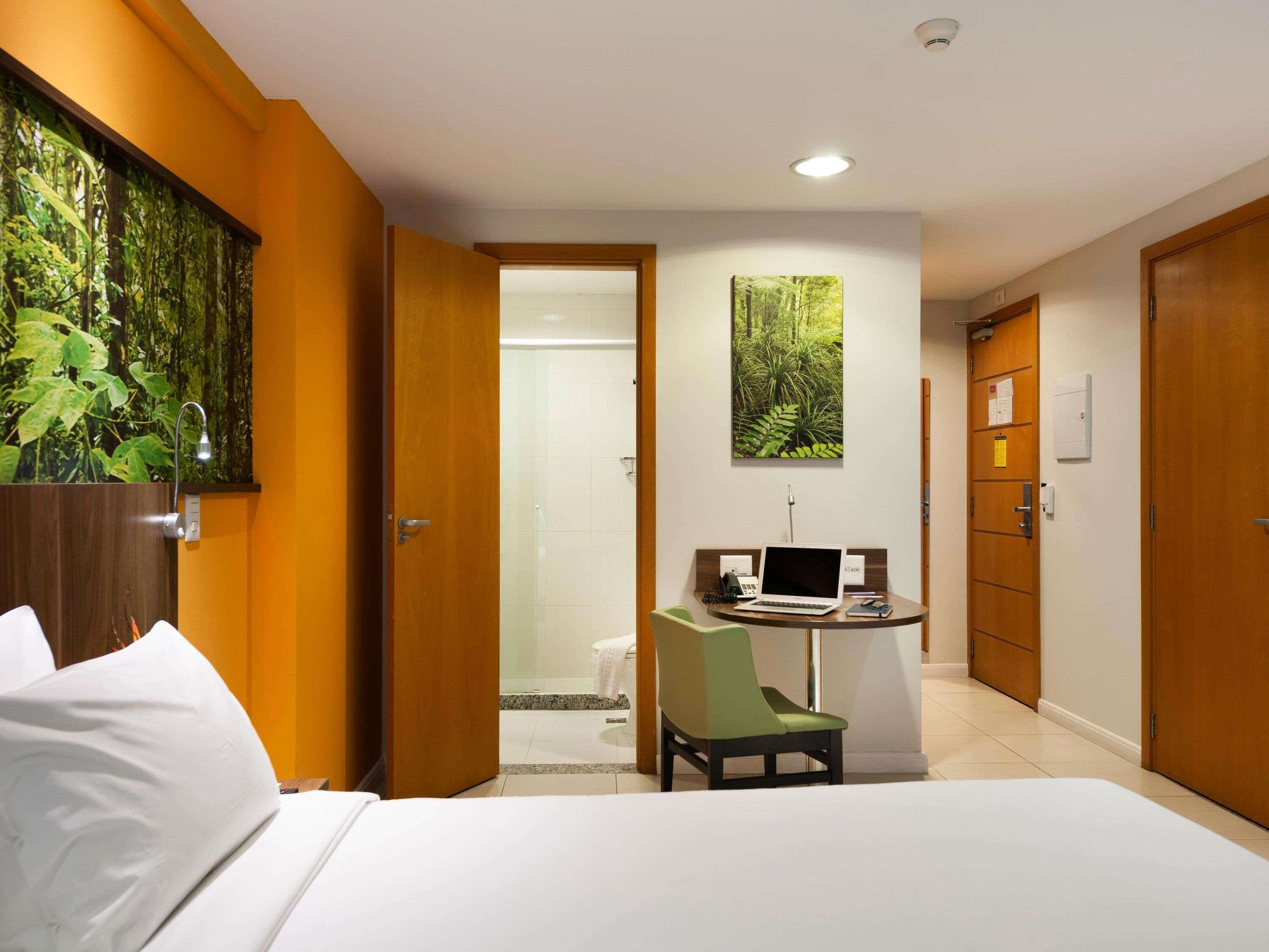 A hotel room with a bed, a desk with a laptop, a chair, and a shower area.