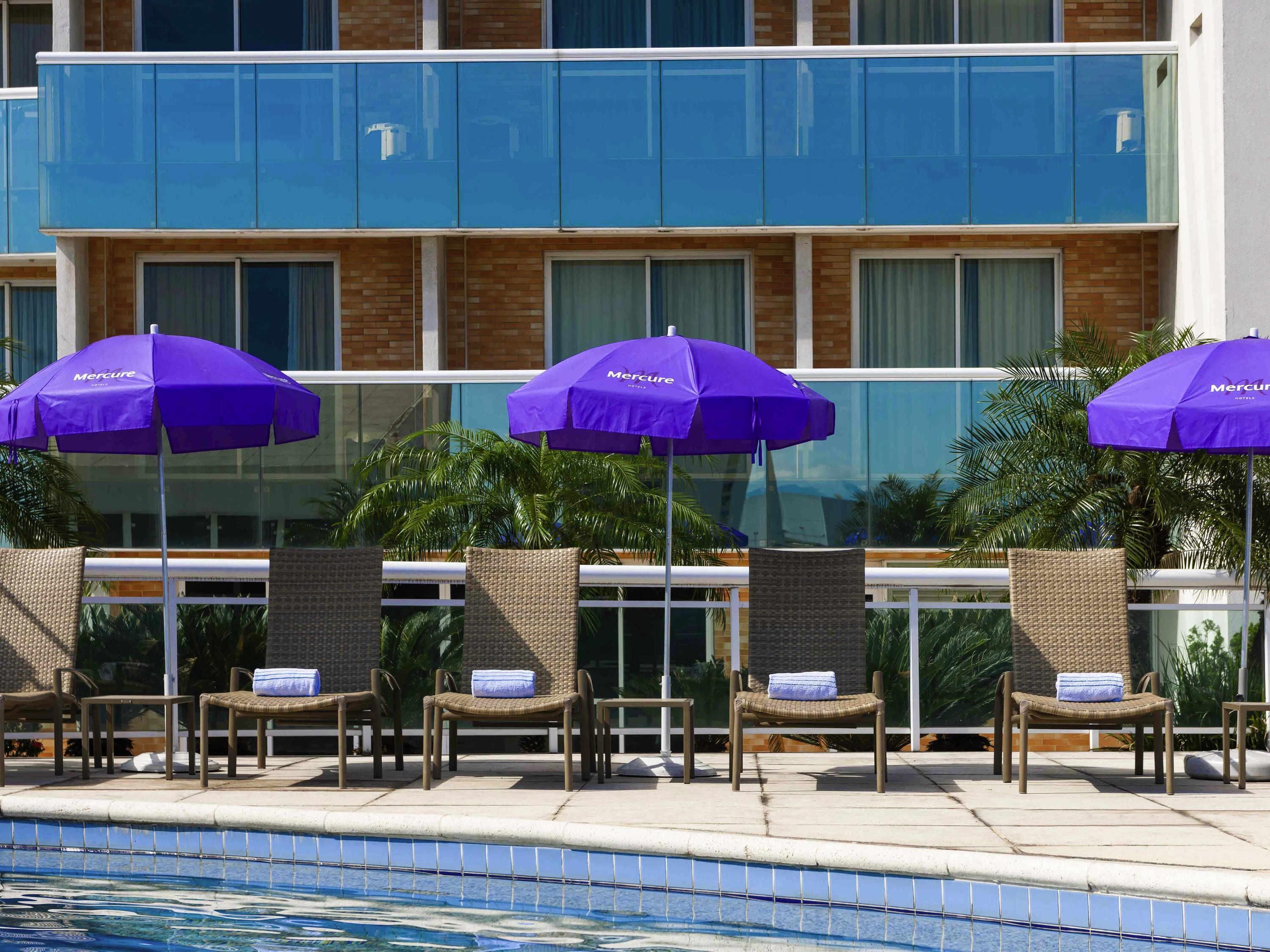 A poolside area with lounge chairs and purple umbrellas, a tiled pool, and a building with large windows and a brick facade.