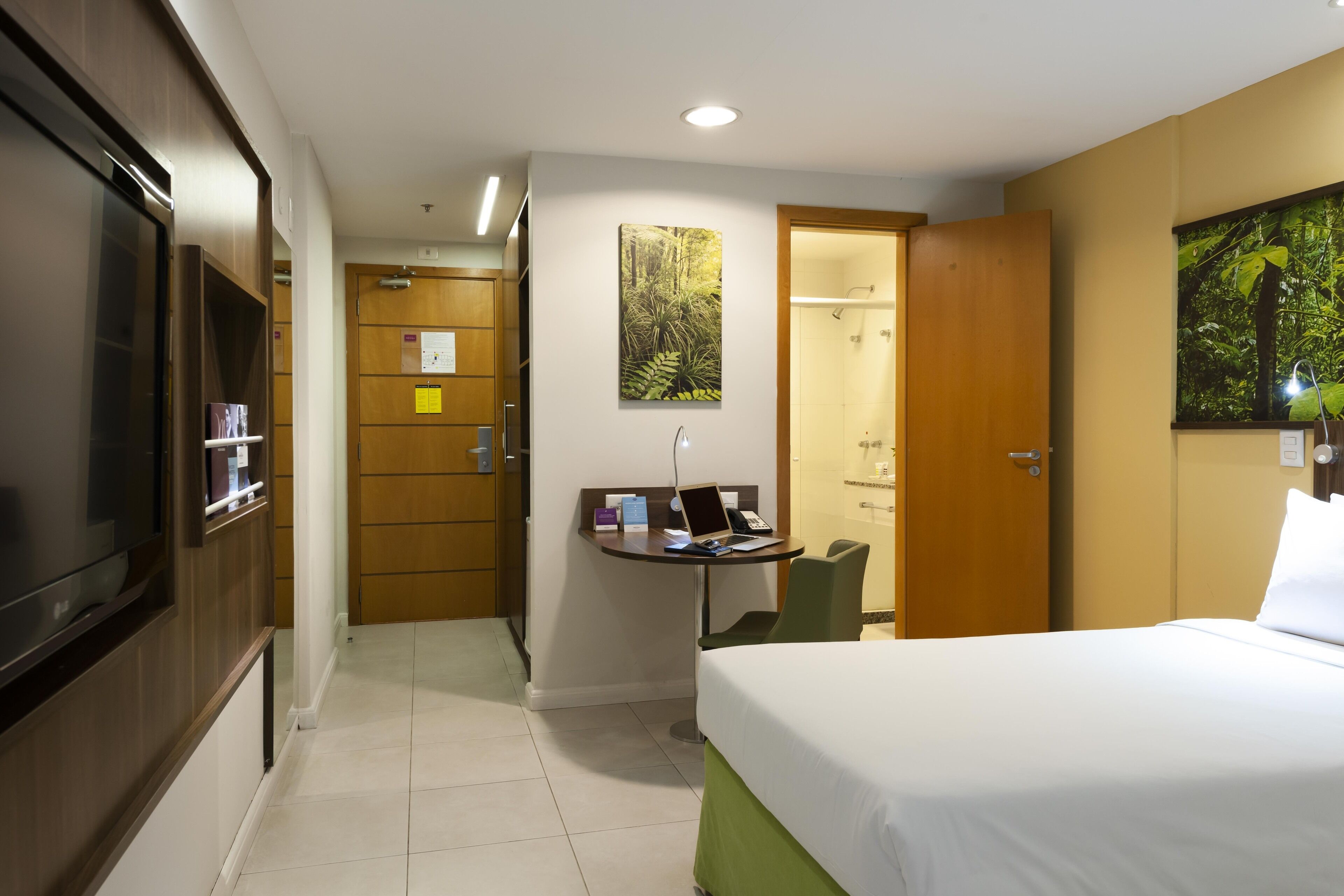 A hotel room with a bed, a desk with a laptop, a television, and a shower area.