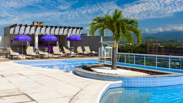 A swimming pool with lounge chairs and umbrellas, surrounded by a tiled deck and a palm tree. - Mercure RJ Nova Iguaçu Hotel (Nova Iguaçu)