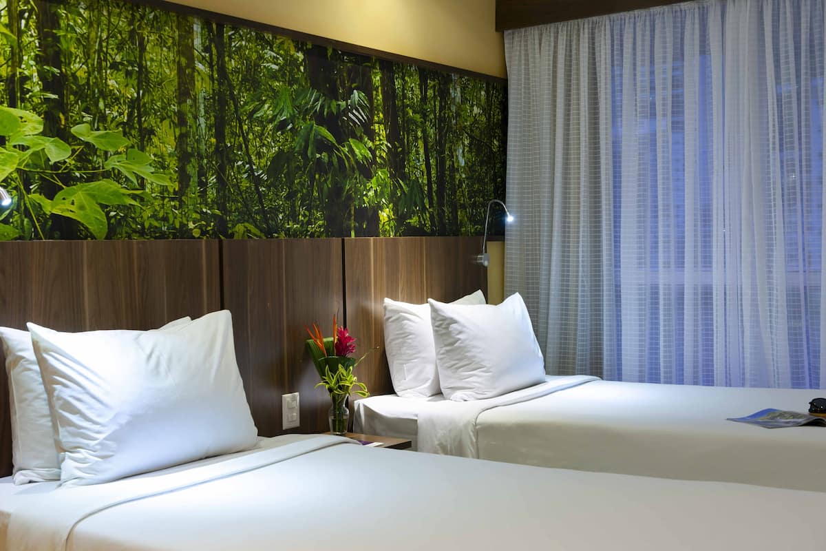 A hotel room with two beds, a large wall mural of a forest, a nightstand with a lamp, and a window with sheer curtains.