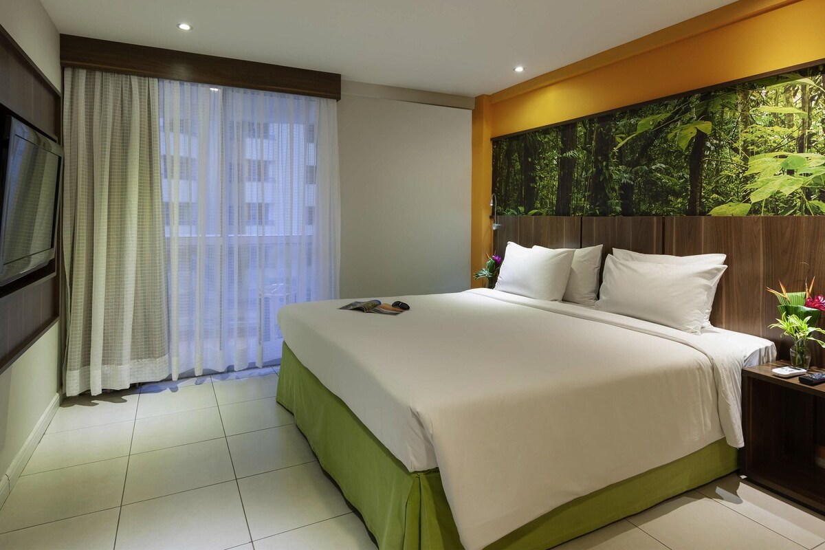 A hotel room with a bed, a TV mounted on the wall, a window with sheer curtains, and a wall mural of a forest.