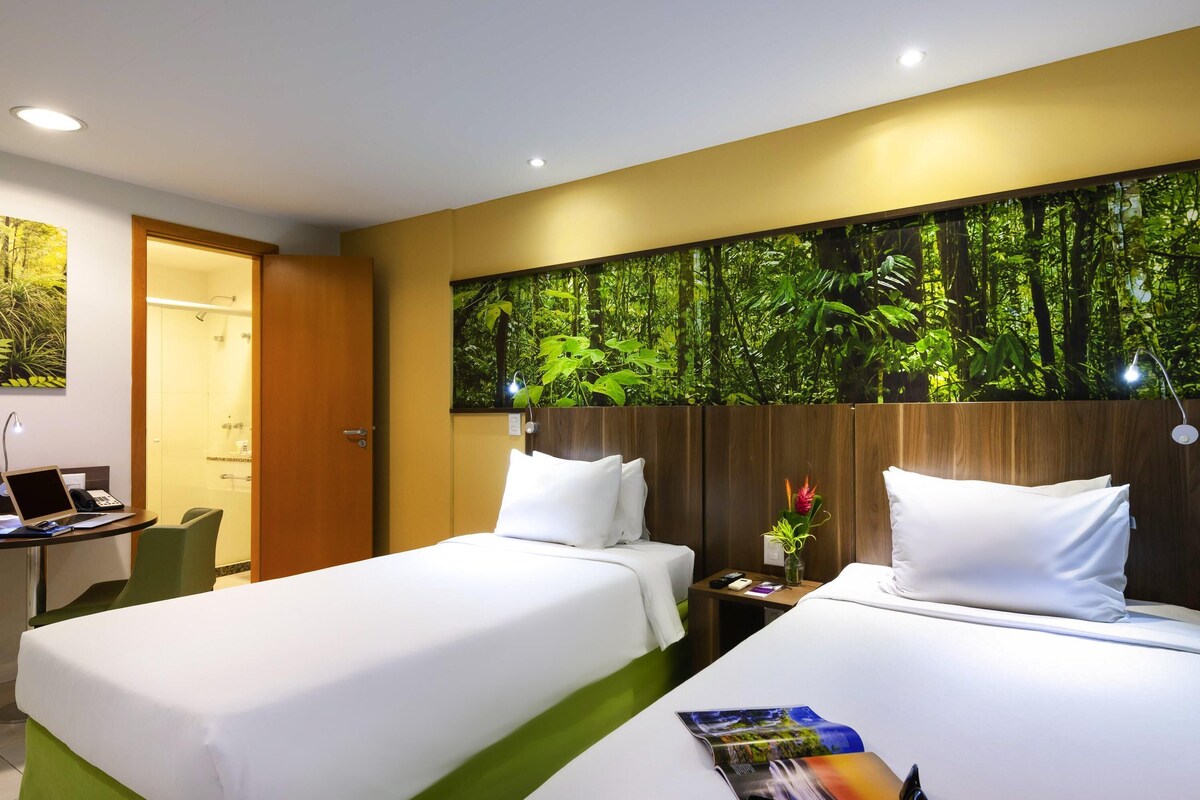 A hotel room with two beds, a desk with a laptop, a shower, and a wall with a forest mural.