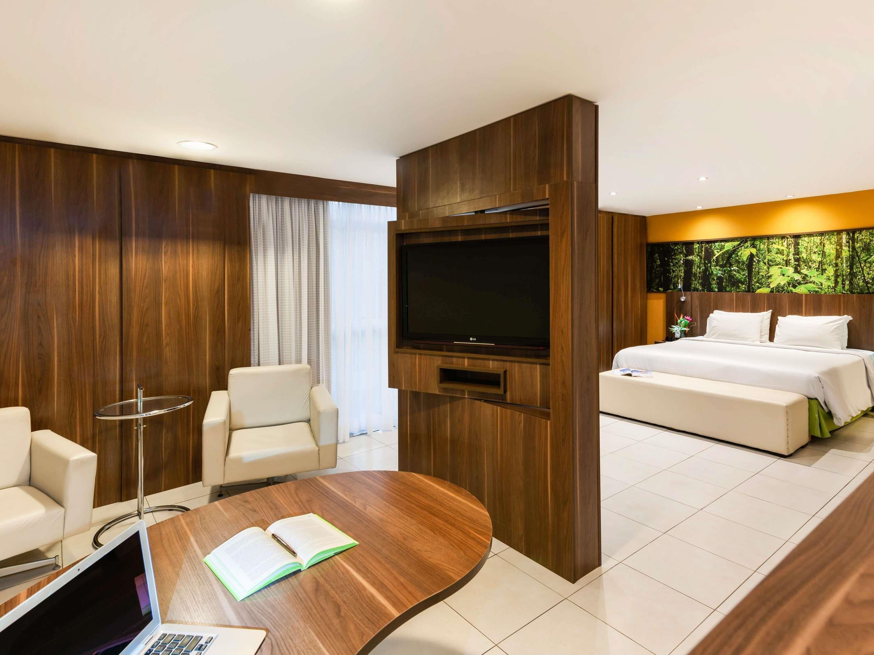 A modern hotel room with a wooden accent wall, a flat-screen TV, a bed, a seating area with a table, and a laptop.