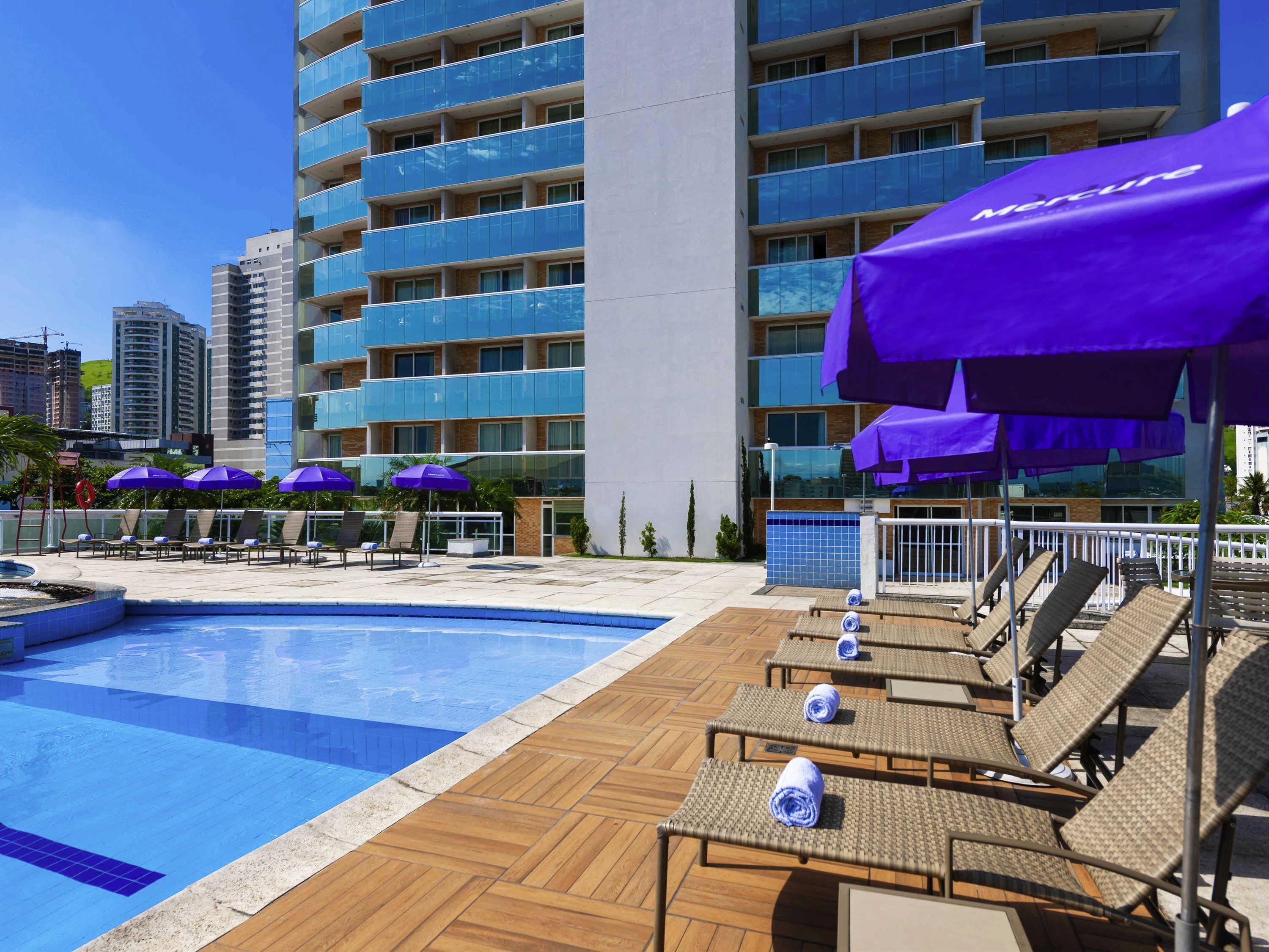 Área de piscina de hotel com espreguiçadeiras e guarda-sóis, uma piscina azul cristalina e um edifício moderno ao fundo.
