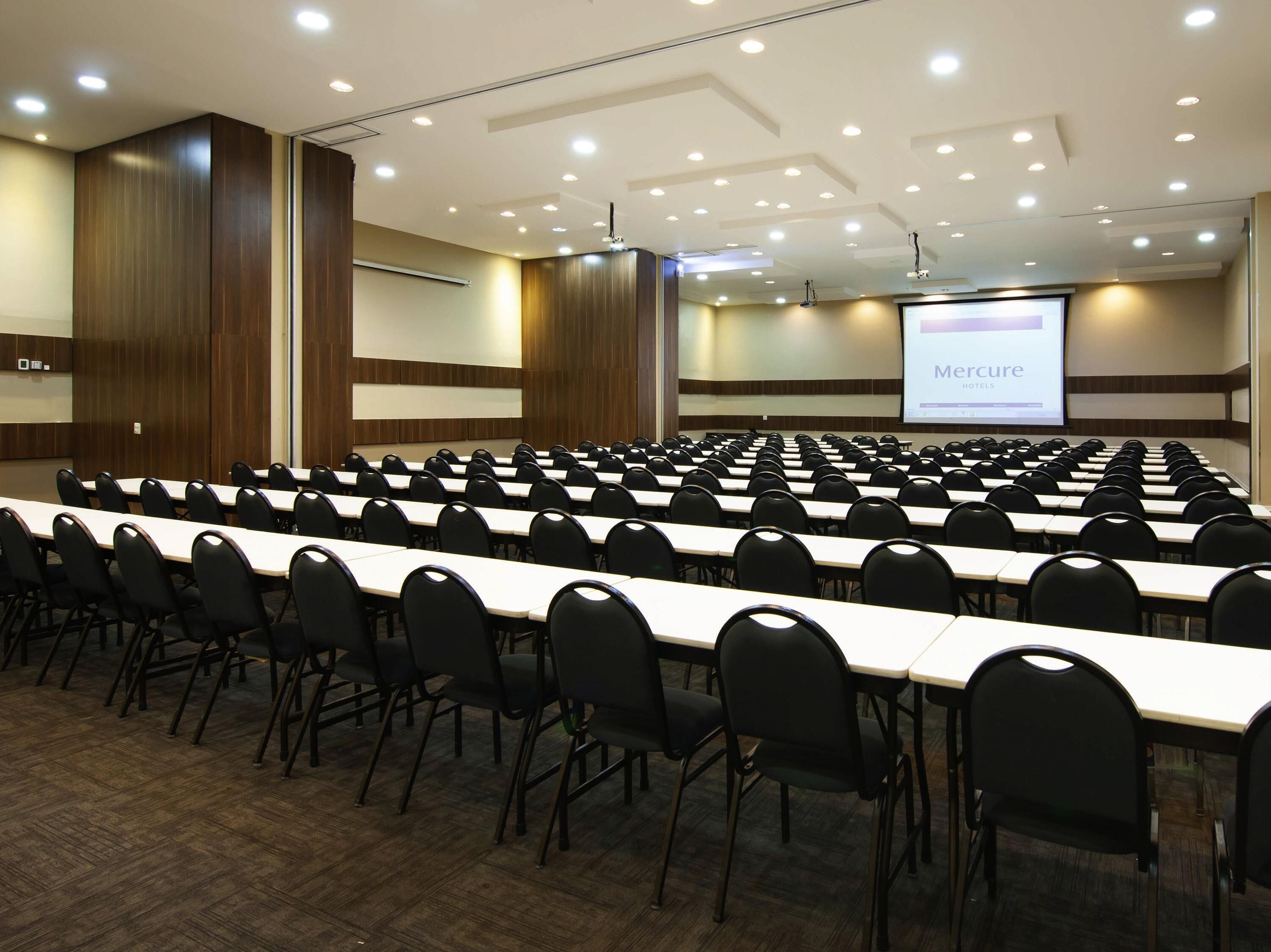 A conference room with rows of tables and chairs, a projection screen, and a wooden panel wall.