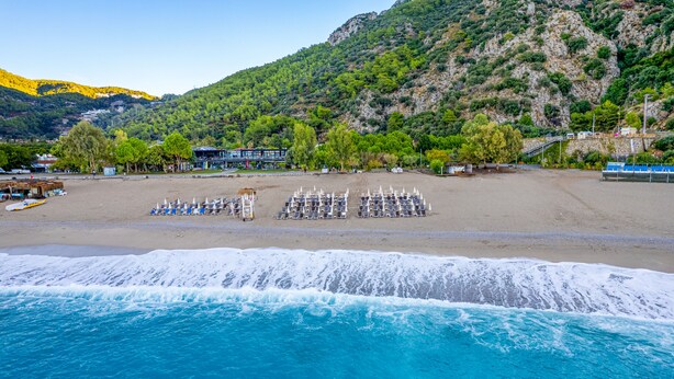 Ein Strand mit Liegestühlen und Sonnenschirmen, ein Gebäude im modernen Stil und eine Bergkulisse im Hintergrund.