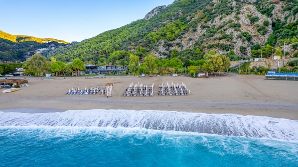 A beach with rows of sun loungers and umbrellas, a building with a modern design, and a mountainous backdrop.