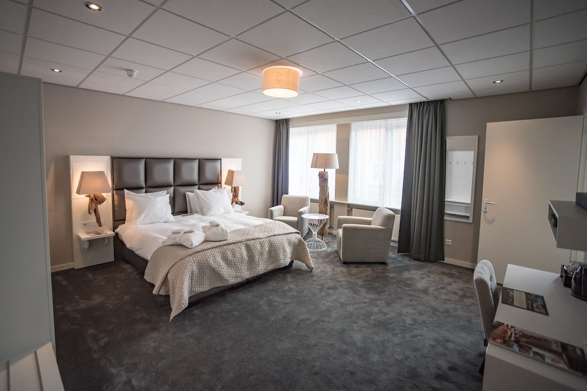 A hotel room with a large bed, two bedside tables with lamps, a small round table, a chair, and a desk with a chair.