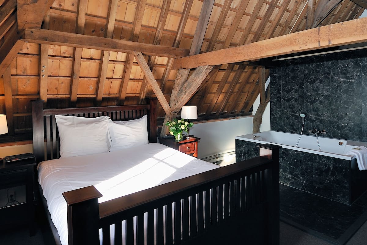 A bedroom with a wooden ceiling, a bed with white bedding, a nightstand, a bedside table with a lamp, and a bathroom with a marble sink.