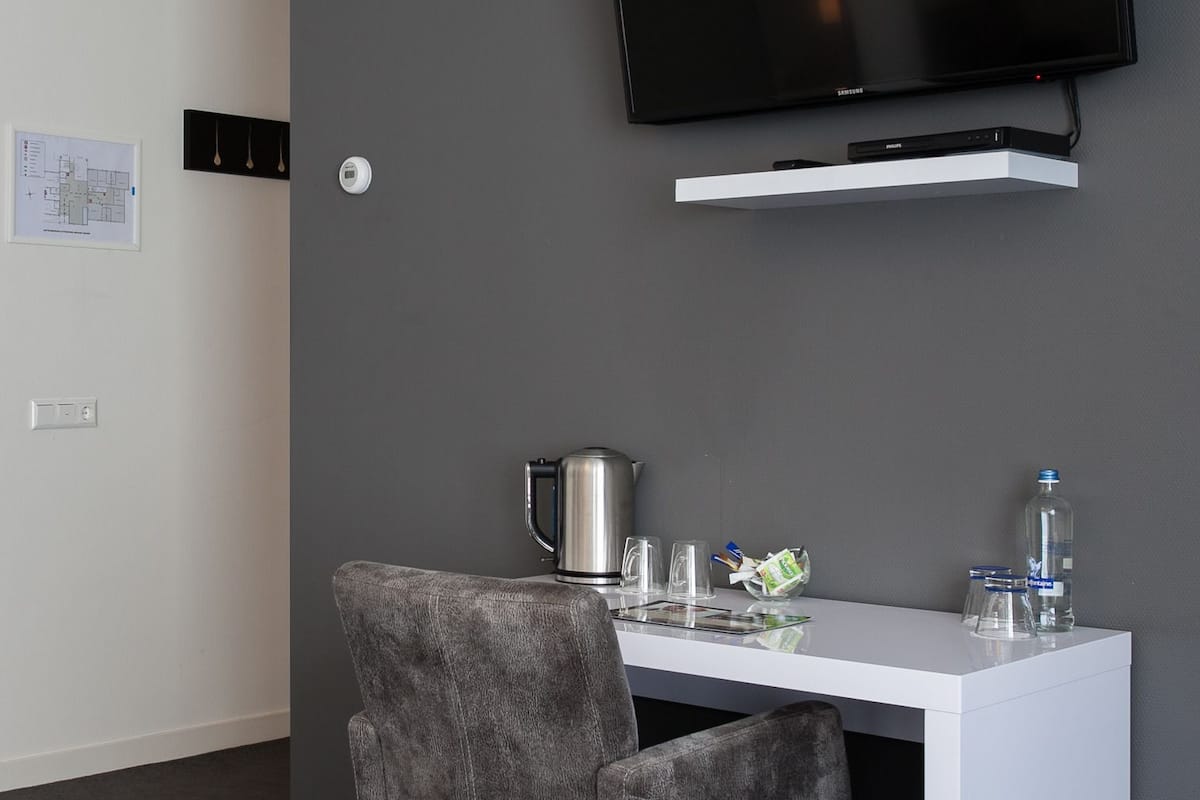 A modern office space with a white desk, a grey upholstered chair, a coffee maker, a water bottle, and a mounted flat-screen TV.