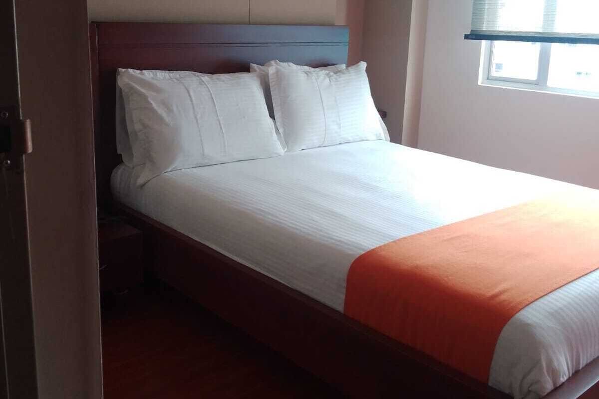 A single bed with white bedding and an orange throw, a wooden headboard, and a window with blinds.