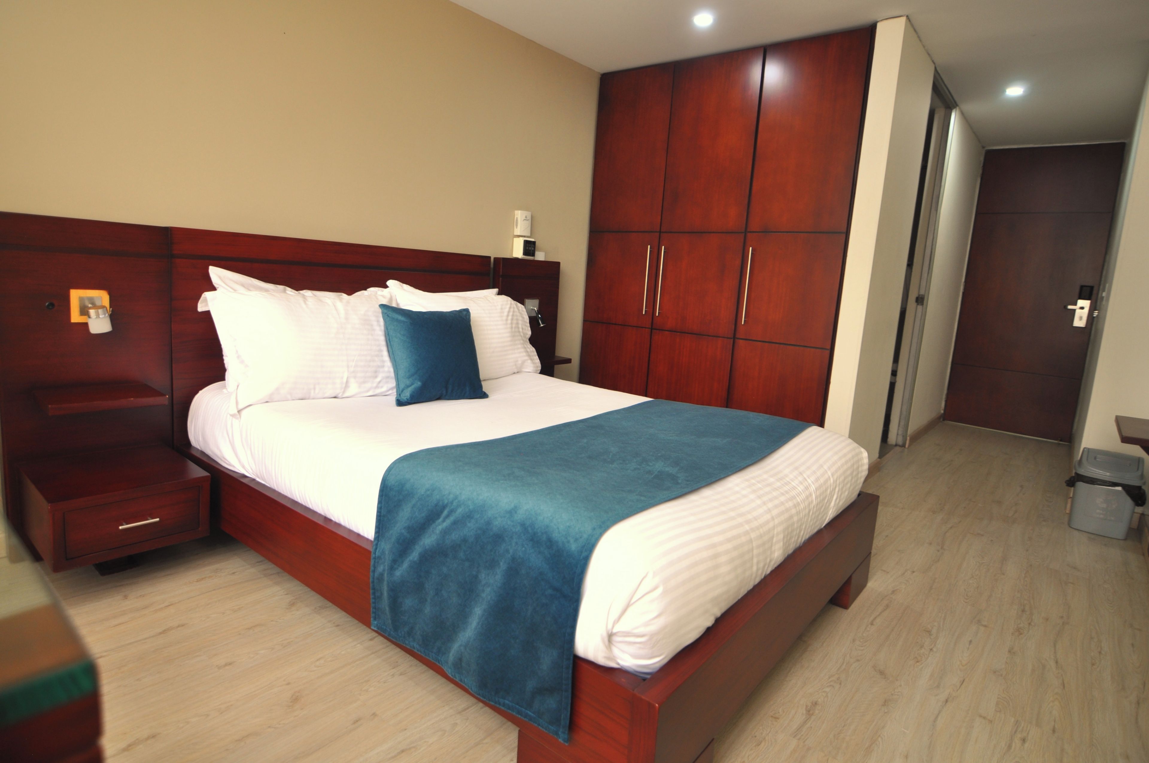 A hotel room with a large bed, wooden wardrobe, bedside tables, and a trash bin.