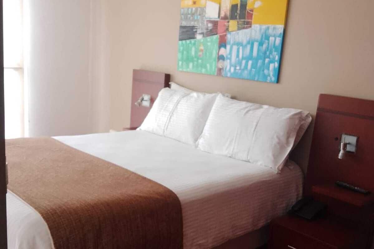 A hotel room with a bed, a nightstand, a bedside table, and a painting on the wall.