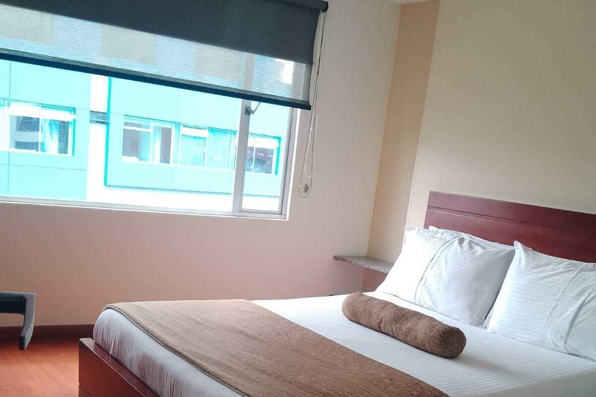A hotel room with a large bed, a wooden headboard, a window with a view of buildings, and a bedside table.