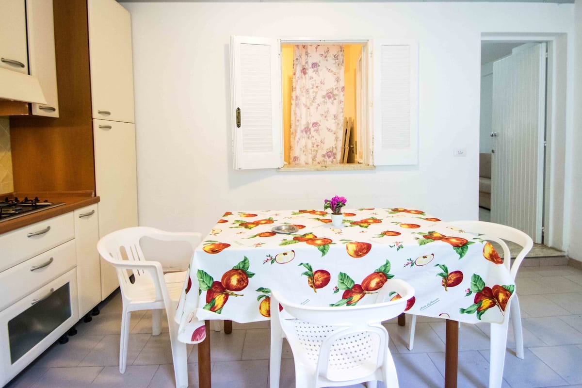 A dining area with a table covered by a fruit-patterned tablecloth, surrounded by white chairs.