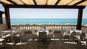 A covered outdoor seating area with tables and chairs, overlooking a beach with clear blue water and boats. - Resort Hotel Rocca di Vadaro (Ricadi)
