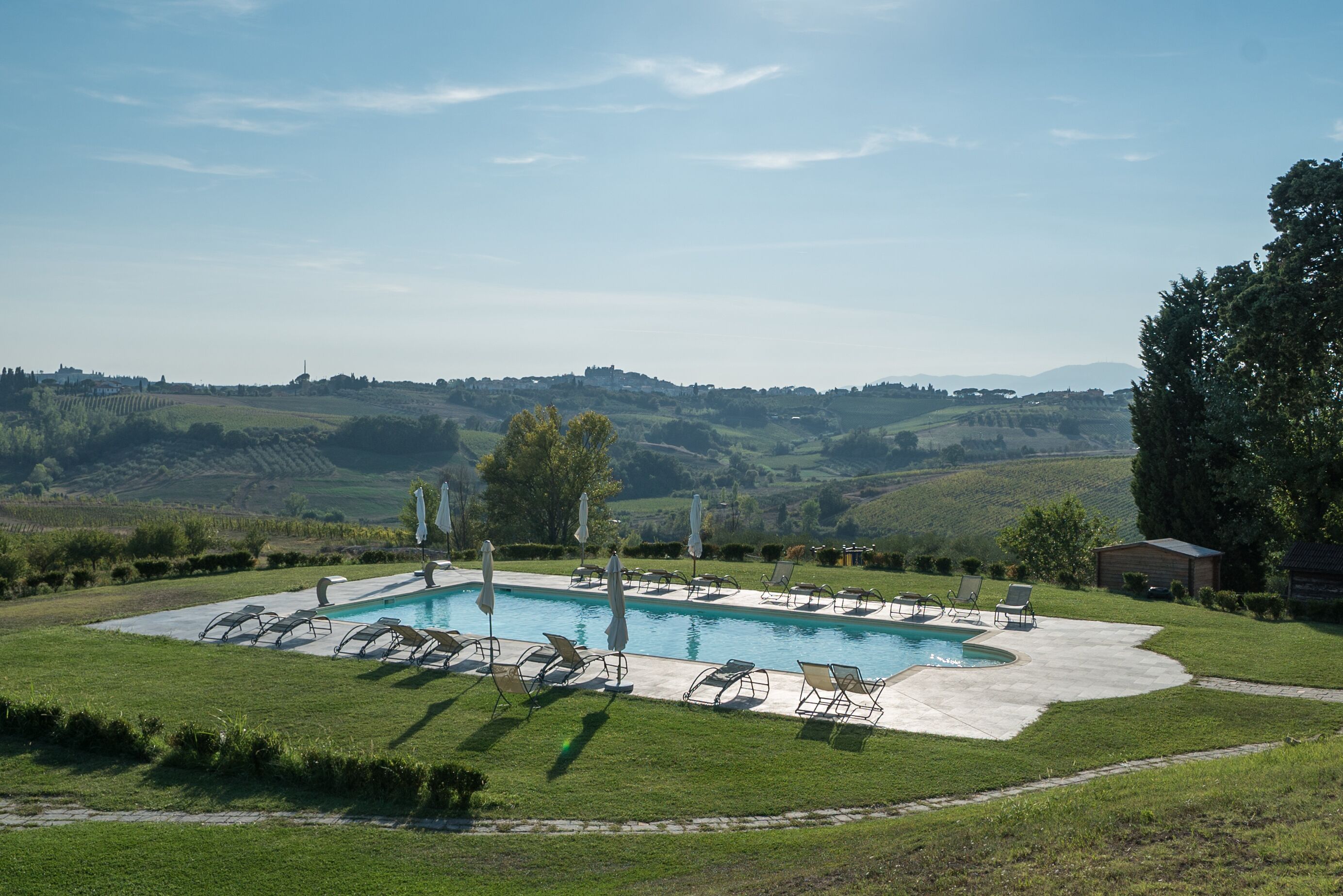 A swimming pool with lounge chairs and umbrellas, surrounded by a grassy area and hills in the background.