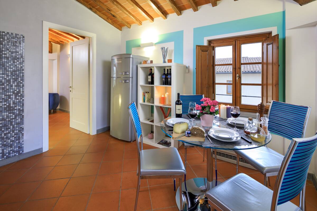 Un comedor con una mesa de vidrio, sillas, un refrigerador y una estantería con botellas.