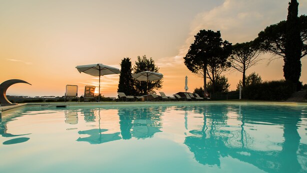 Una piscina con sillas de descanso y sombrillas, rodeada de árboles y un cielo al atardecer.