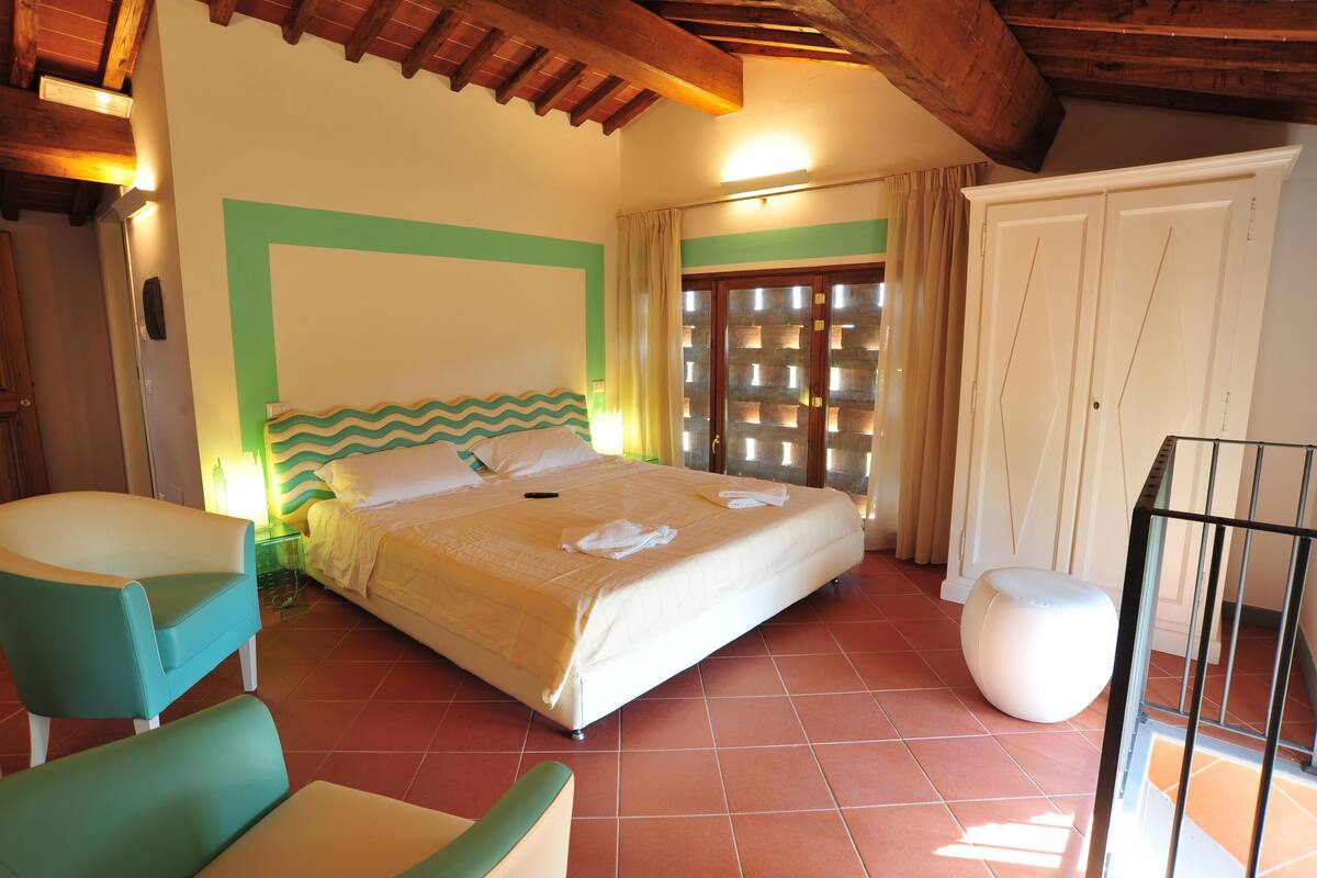 Un dormitorio con cama, una silla, un taburete pequeño, un armario y una ventana con cortinas.