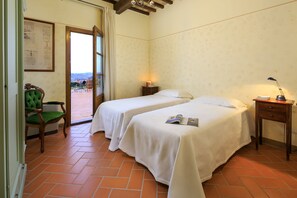 Une chambre d’hôtel avec deux lits, un fauteuil vert, une petite table avec une lampe et une vue sur l’extérieur, visible à travers une porte ouverte.
