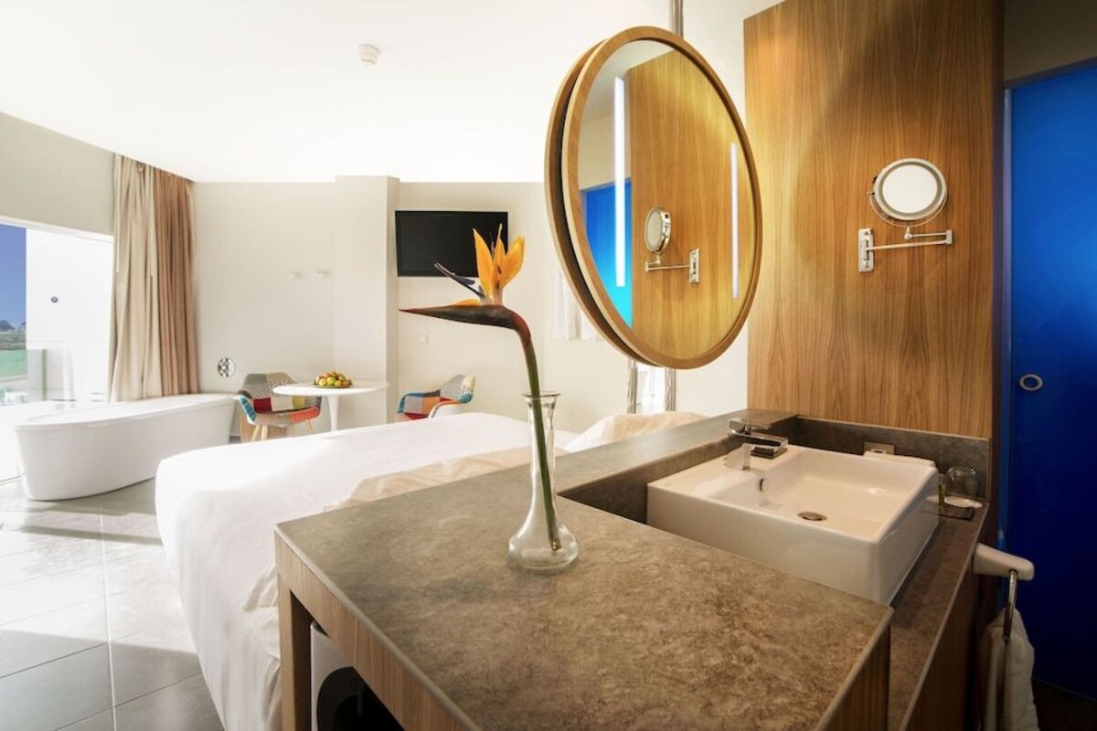 A modern hotel room with a large bed, a bathroom with a bathtub, a round mirror, and a vase with a flower.