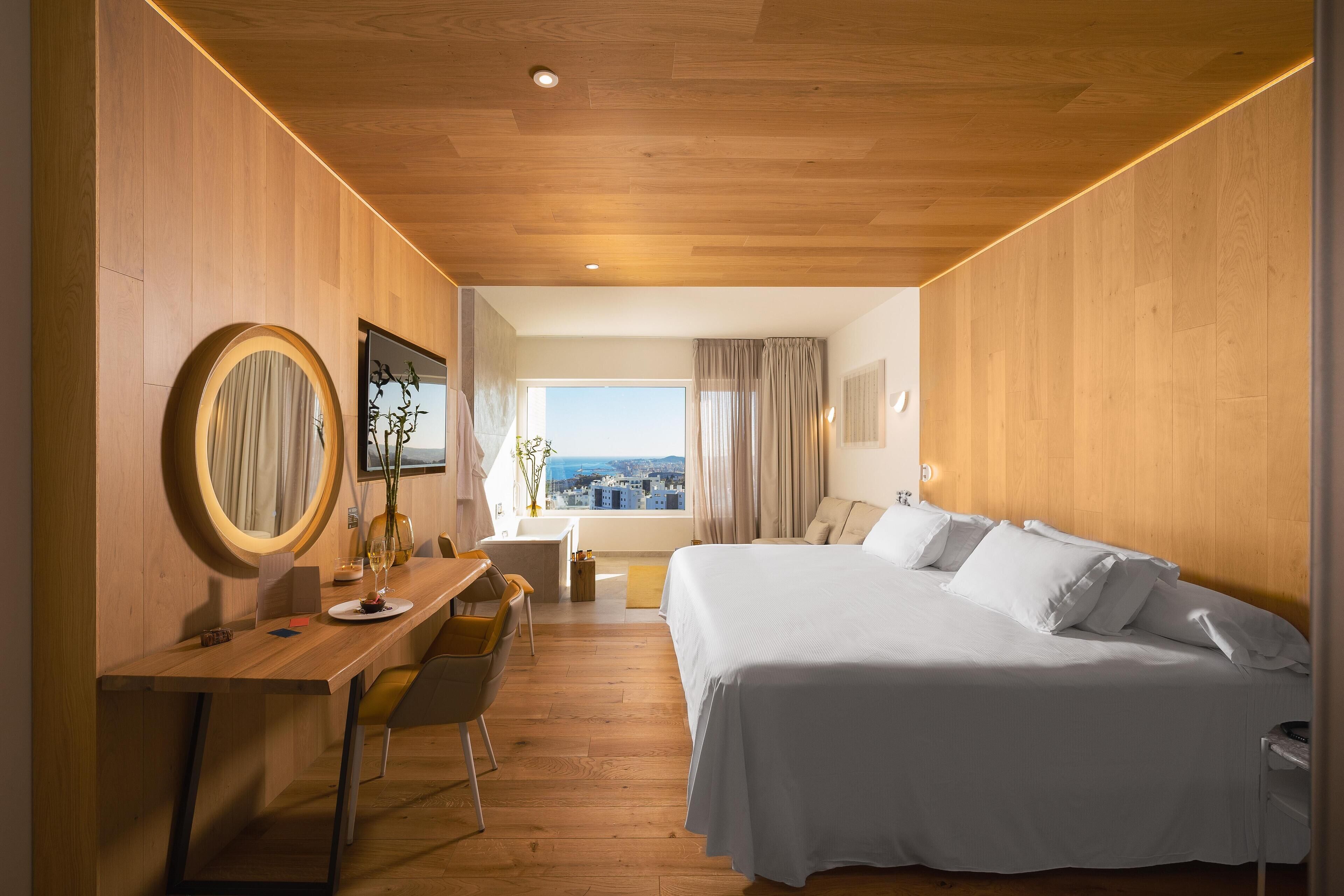 Habitación de hotel con techo de madera, una cama grande, un escritorio con silla, un espejo redondo y vista a la ciudad a través de una ventana.