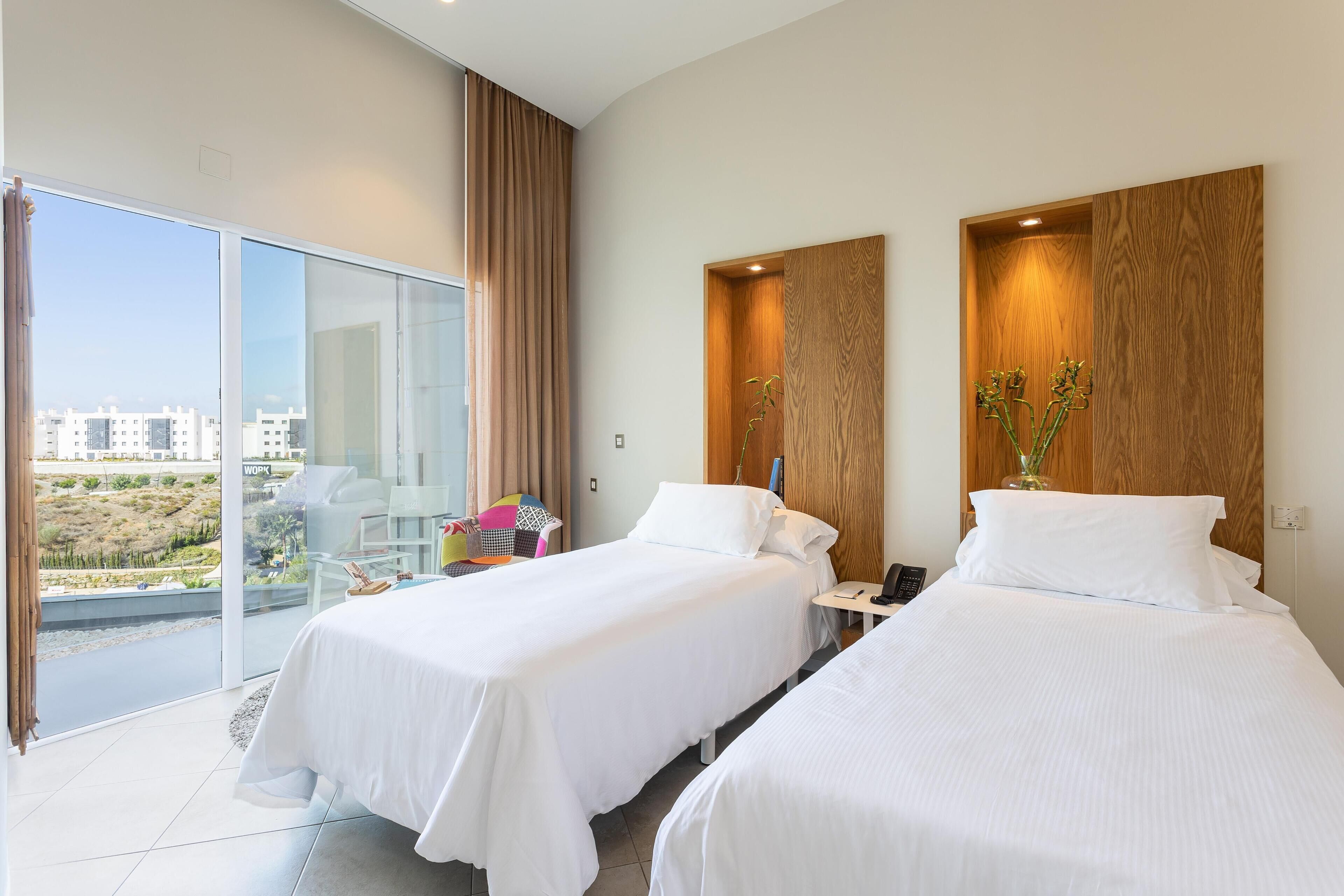 A hotel room with two beds, a balcony with a view of the city, and a wooden accent wall.